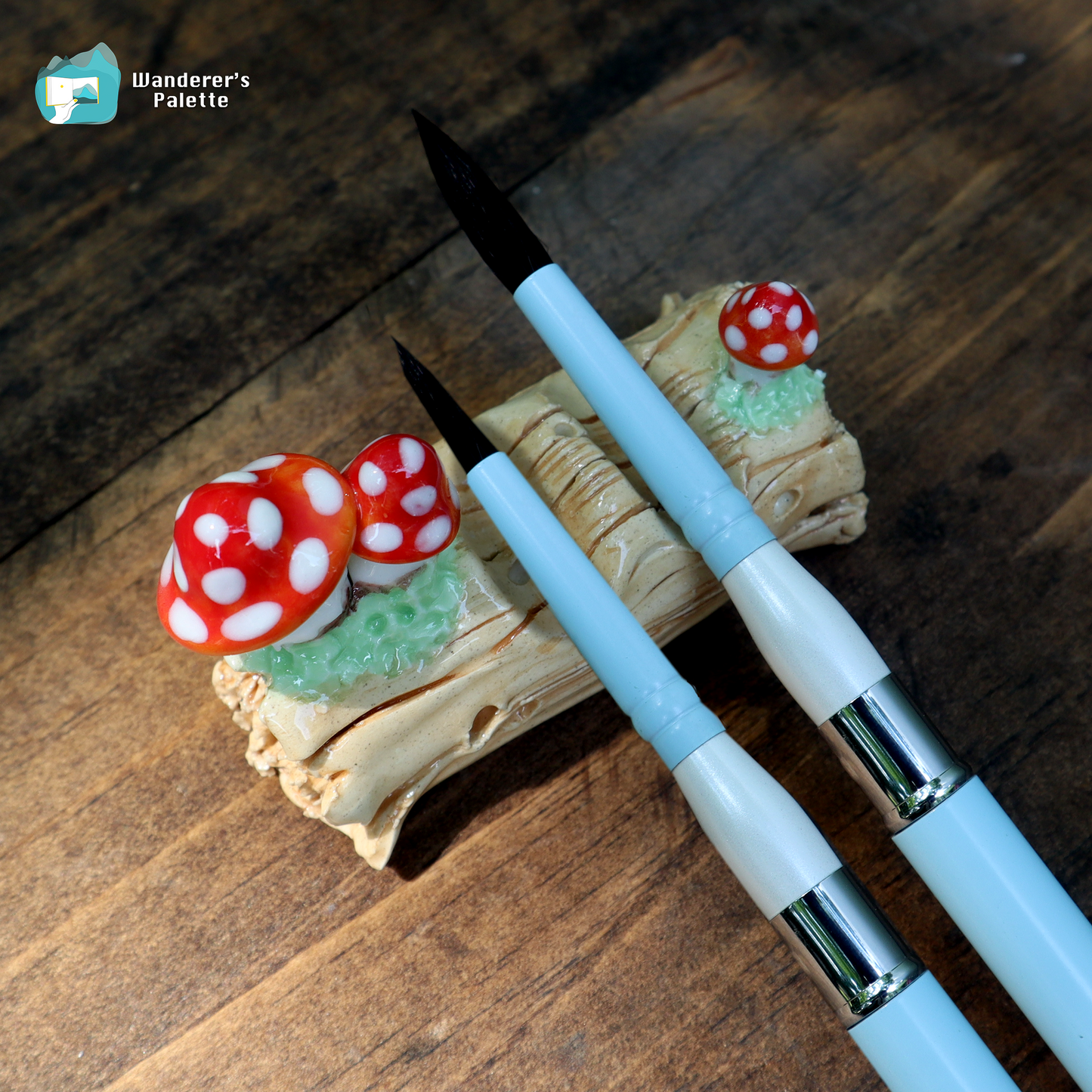 Two light blue and white calligraphy brushes with black bristles are displayed on a decorative wooden stand featuring red and white mushrooms, set against a dark wood background. A 'Wanderer's Palette' logo is visible in the top left corner.