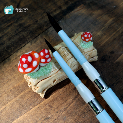 Two light blue and white calligraphy brushes with black bristles are displayed on a decorative wooden stand featuring red and white mushrooms, set against a dark wood background. A 'Wanderer's Palette' logo is visible in the top left corner.