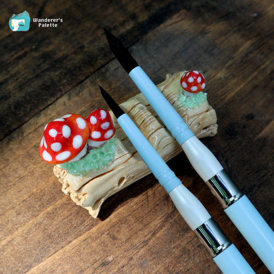 Two light blue and white calligraphy brushes with black bristles are displayed on a decorative wooden stand featuring red and white mushrooms, set against a dark wood background. A 'Wanderer's Palette' logo is visible in the top left corner.