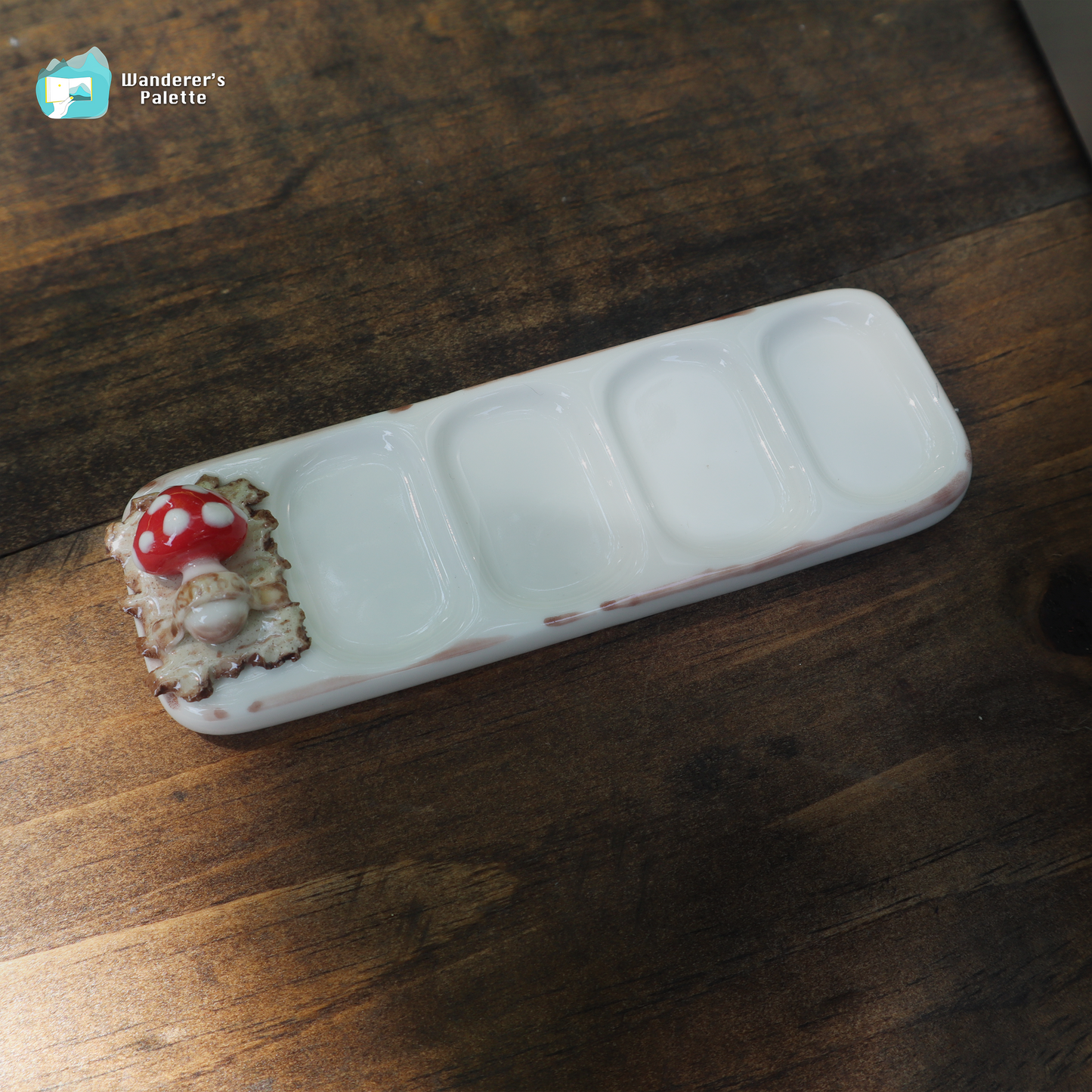 A white ceramic five-compartment palette tray with a rustic brown painted edge and a decorative red-and-white mushroom cap on one end, shown on a dark wooden tabletop. A small 'Wanderer's Palette' logo watermark appears in the top-left corner.