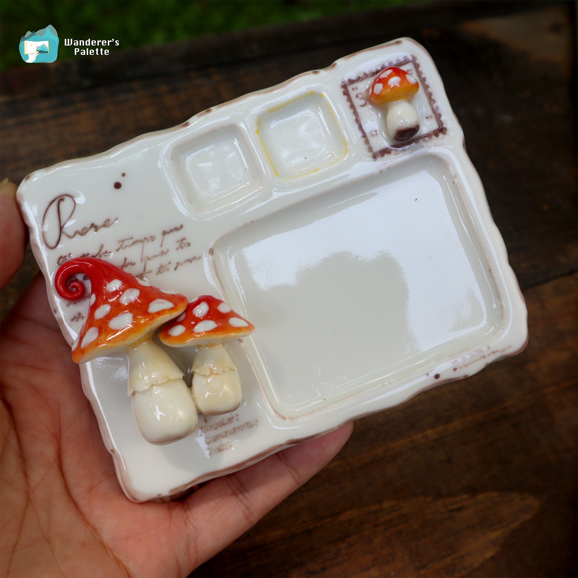 A hand holds a small white ceramic paint palette with three compartments, a large mixing well, decorative mushroom motifs, and the word 'Piero' printed on the side. The background is a dark wooden surface.