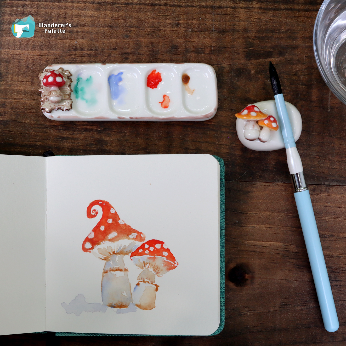 An open sketchbook on a dark wooden table showing a painted pair of red-and-white mushrooms. Surrounding the sketchbook are a white paint palette with mixed paint colors, a small mushroom-shaped paint holder, a light blue paintbrush, and part of a glass of water.