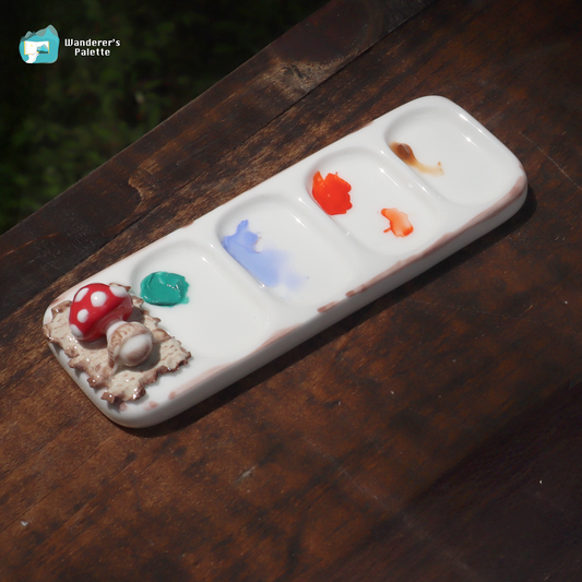 A white ceramic artist palette with five compartments, decorated with a mushroom and shell motif on one end, resting on a dark wooden surface. The palette is shown with various paint colors in the wells, including red, green, blue, orange, and brown.
